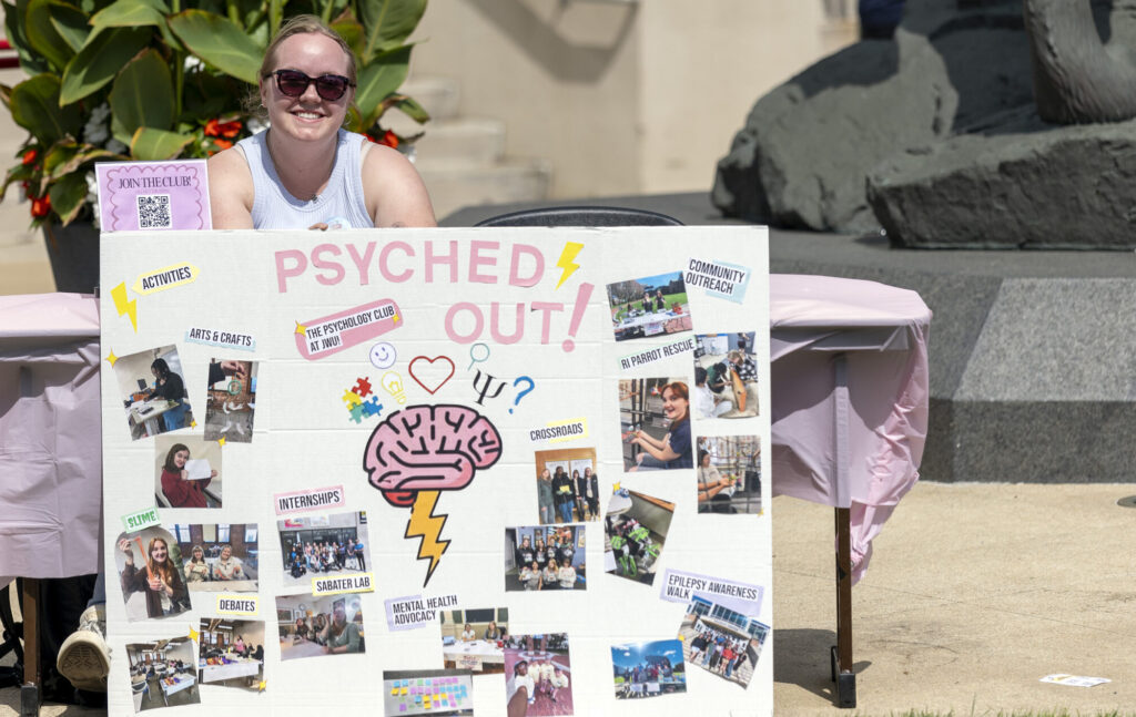 A smiling person stands behind a large promotional board for “Psyched Out!” featuring photos and information about mental health activities and clubs.