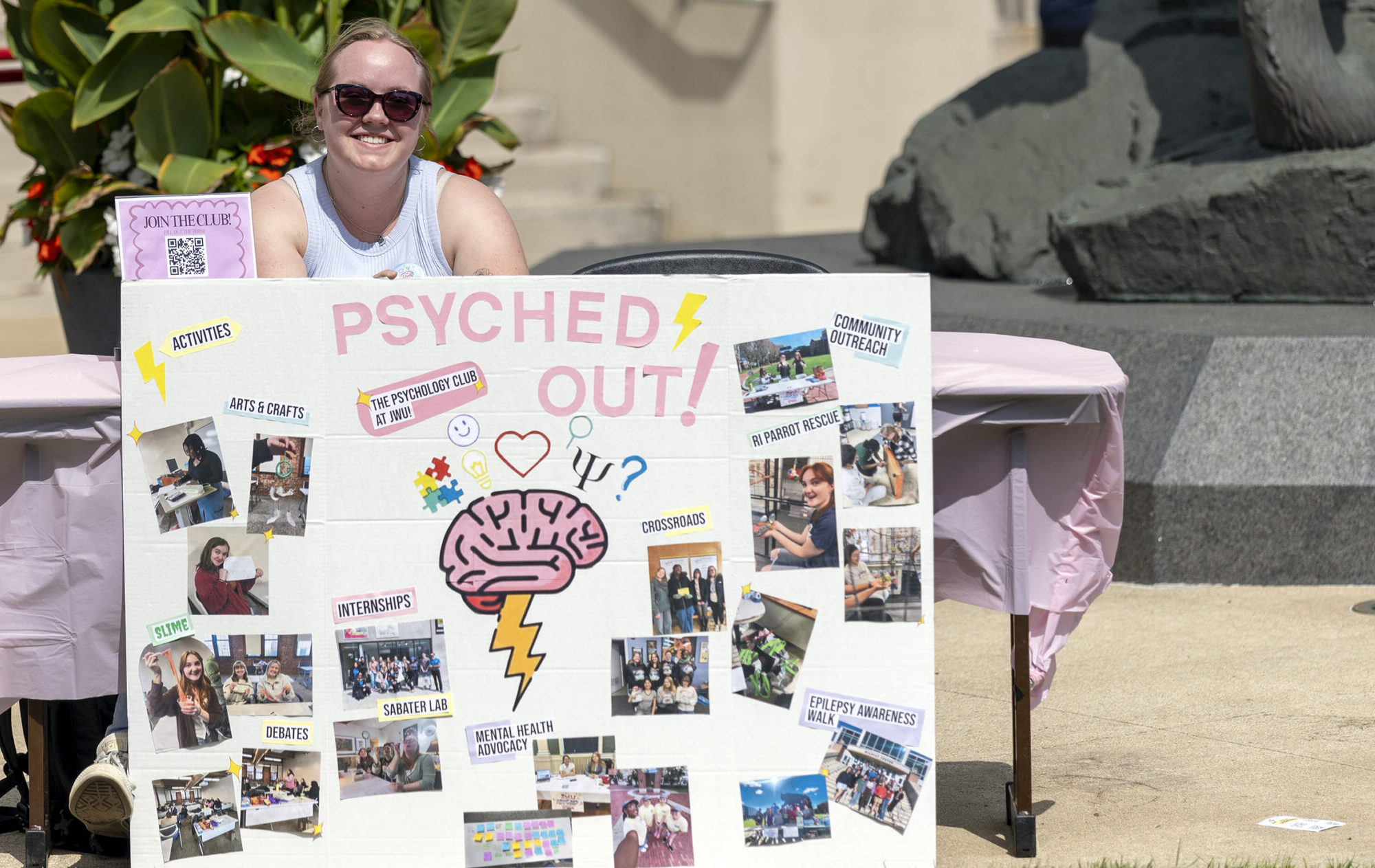 A smiling person stands behind a large promotional board for “Psyched Out!” featuring photos and information about mental health activities and clubs.