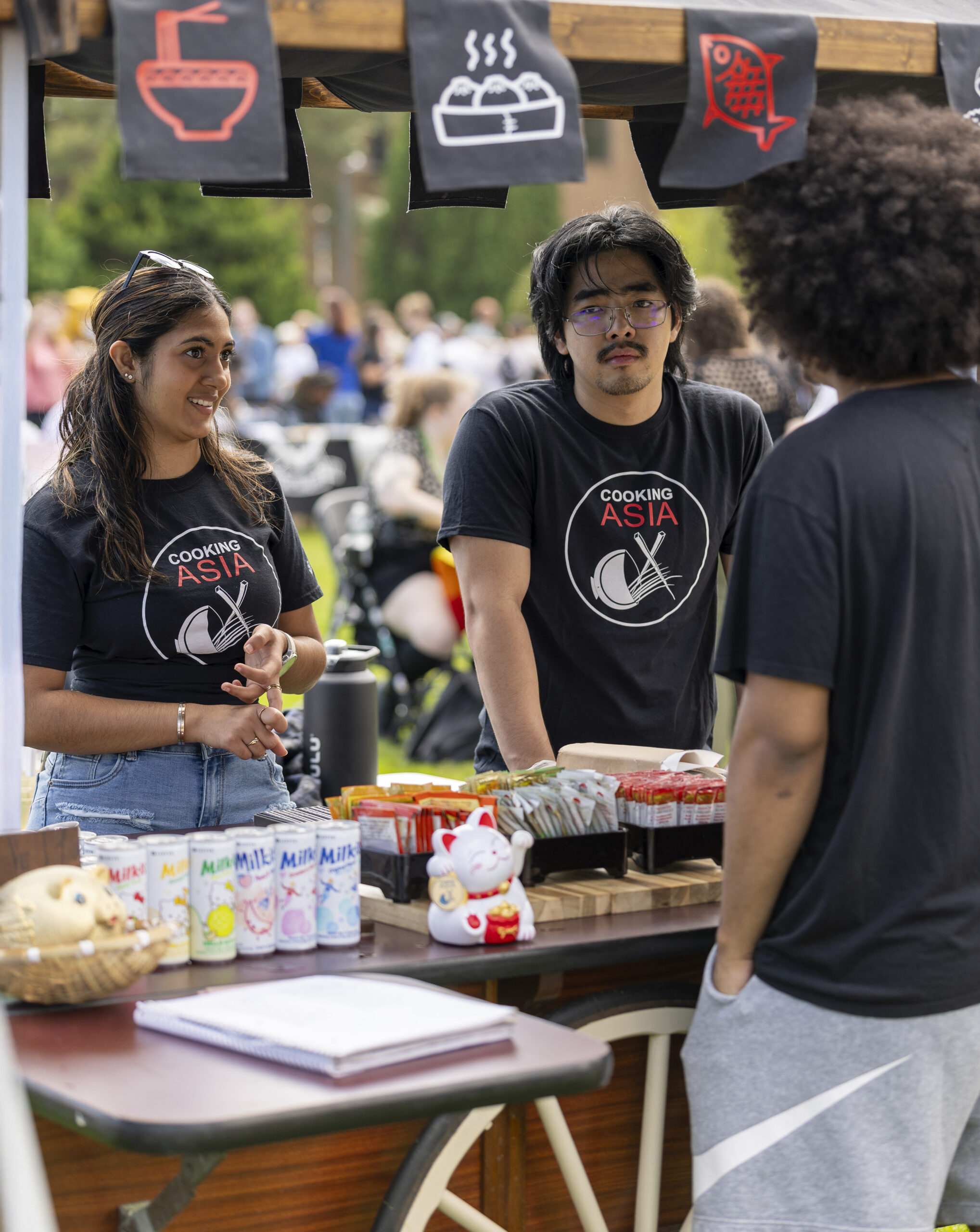 Three individuals wearing "Cooking Asia" shirts stand behind a food stall at an outdoor event, engaging with a customer.