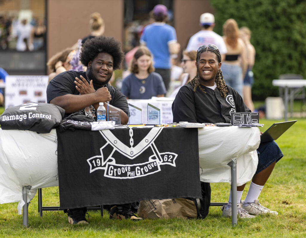 Two individuals sit at an outdoor table displaying merchandise and promotional materials for a group called "Groove Phi Groove."