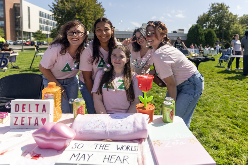 Five individuals in pink Delta Zeta shirts pose cheerfully around a table displaying branded merchandise and decorations at an outdoor event.