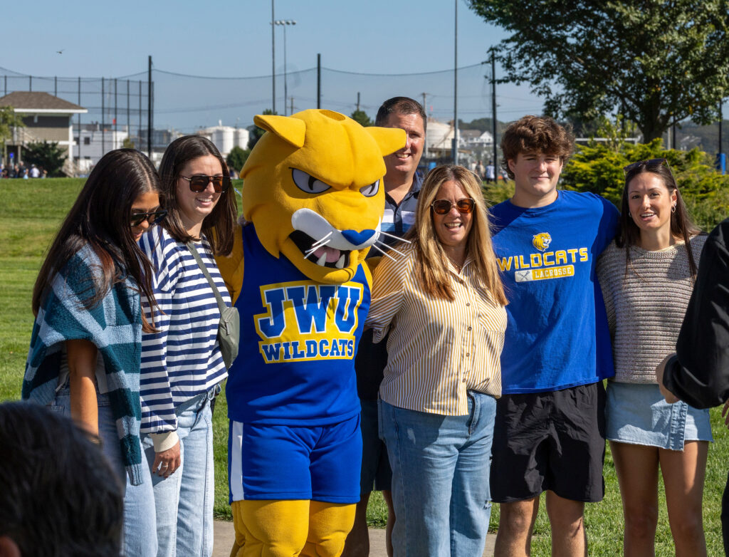 A group of people, including a person in a yellow and blue JWU Wildcats mascot costume, stand together outdoors on a sunny day.