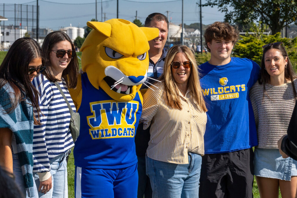 A group of people, including a person in a large yellow and blue mascot costume, stand together outdoors on a sunny day.