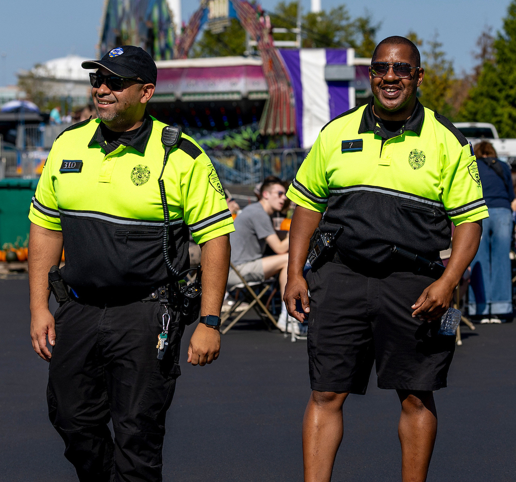 Two officers in bright uniforms walk across a parking lot at an outdoor event with rides and people in the background.