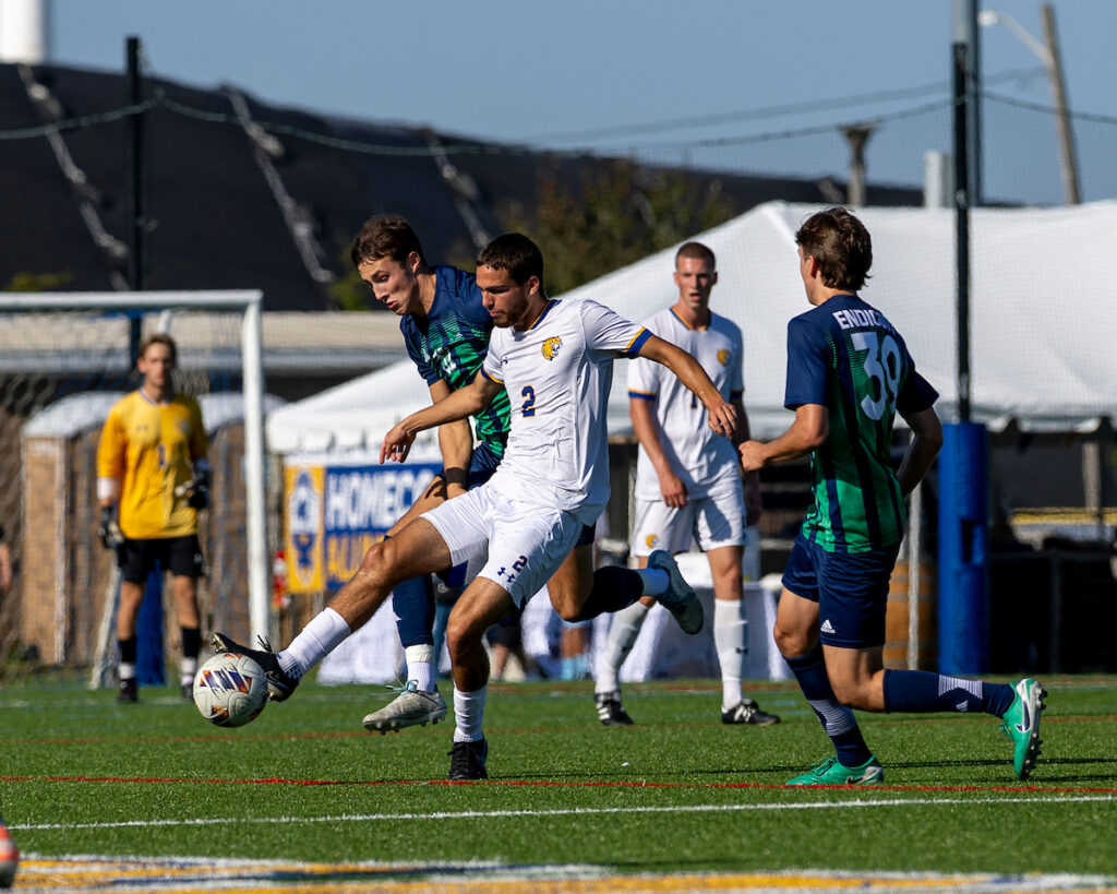 A soccer player in a white jersey controls the ball while being challenged by an opponent in a green and blue uniform during a match.