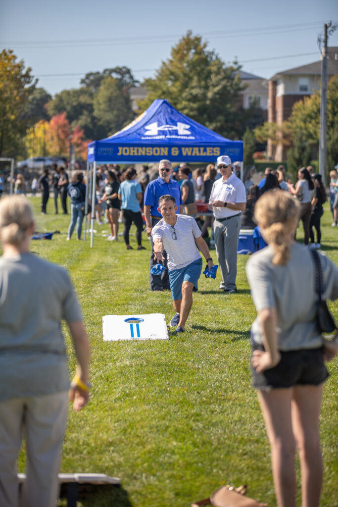 A person throws a disc toward a target on a grassy field at an outdoor event sponsored by Johnson & Wales, with attendees and a branded tent in the background.