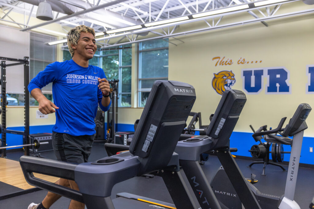A person in a blue "Johnson & Wales Cross Country" shirt smiles while running on a treadmill in a gym with "This is... UR" branding on the wall.