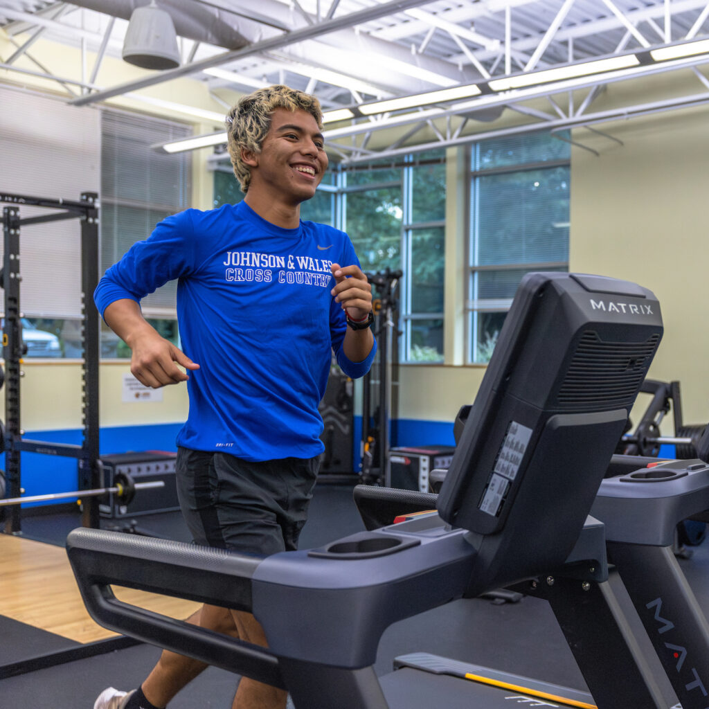 A person in a blue "Johnson & Wales Cross Country" shirt smiles while running on a treadmill in a gym with "This is... UR" branding on the wall.