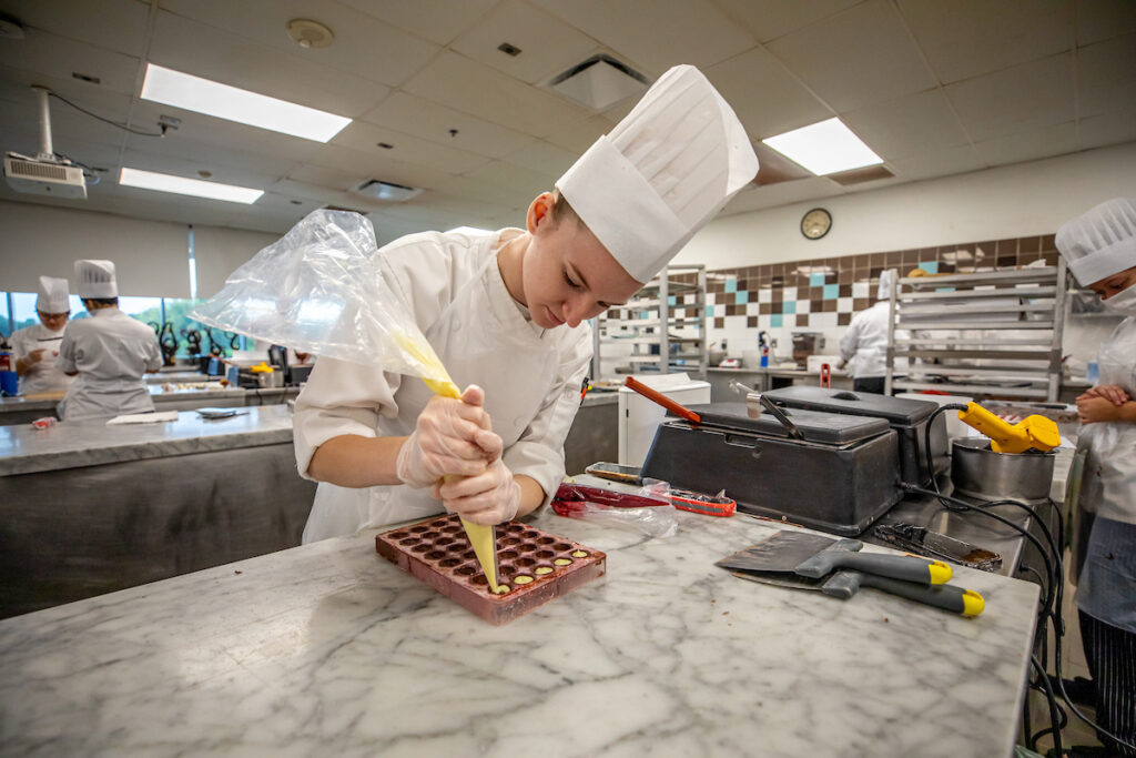 A chef carefully pipes yellow icing into a chocolate mold in a busy professional kitchen.