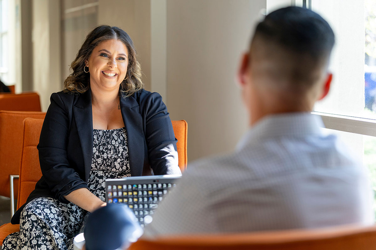 A graduate student in a black blazer and patterned dress smiles warmly while seated across from another individual in a professional setting.
