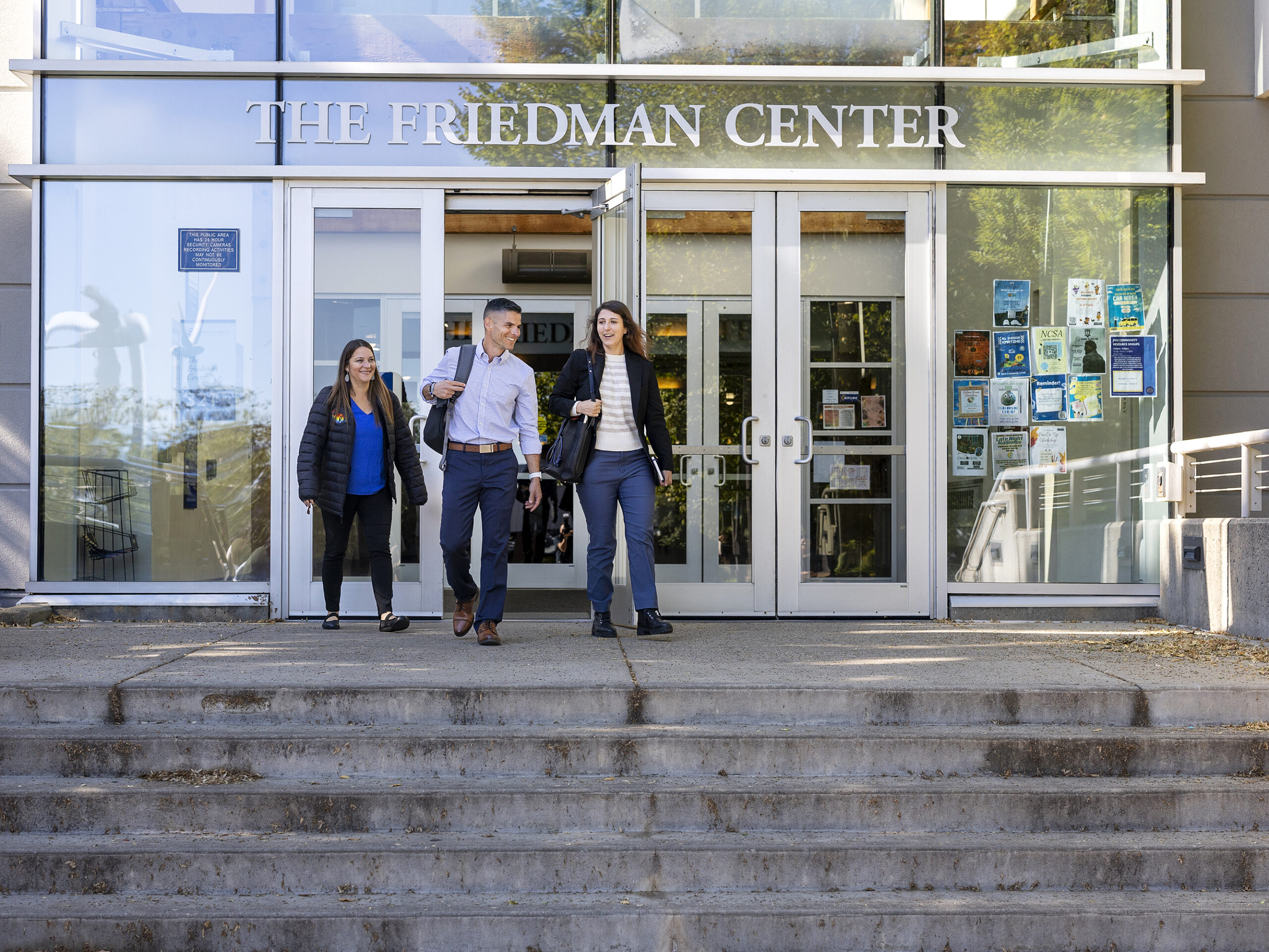 Three people exit The Friedman Center building, walking down steps and talking.
