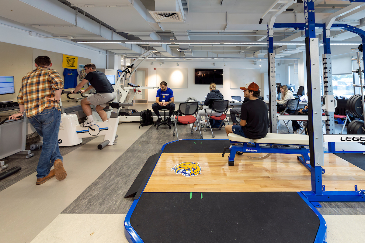 People are working out and using computers in a fitness and training facility with exercise equipment and a logo on the floor.