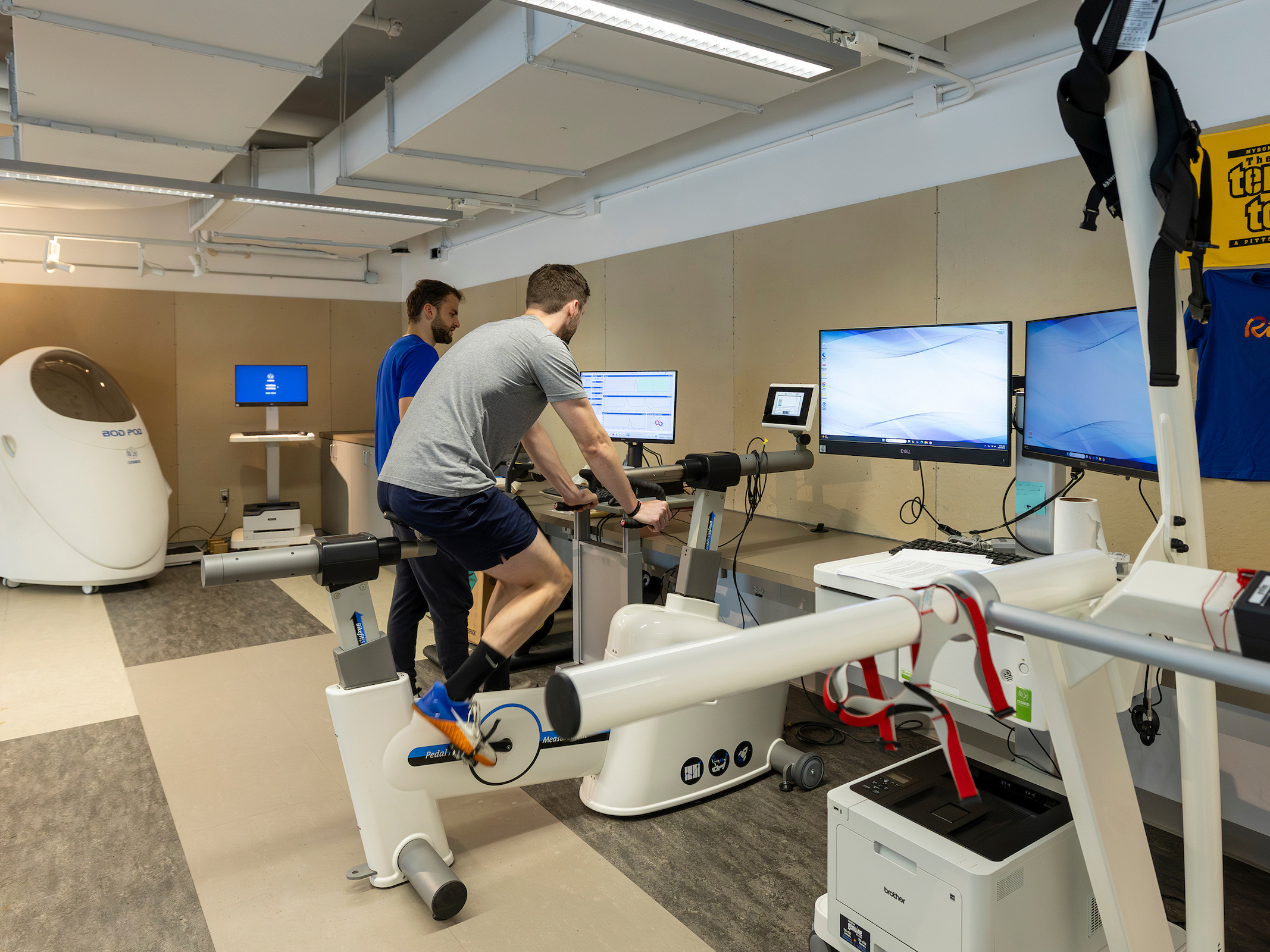 A person cycles on a stationary bike in a lab setting while another observes, with multiple monitors and medical equipment visible.