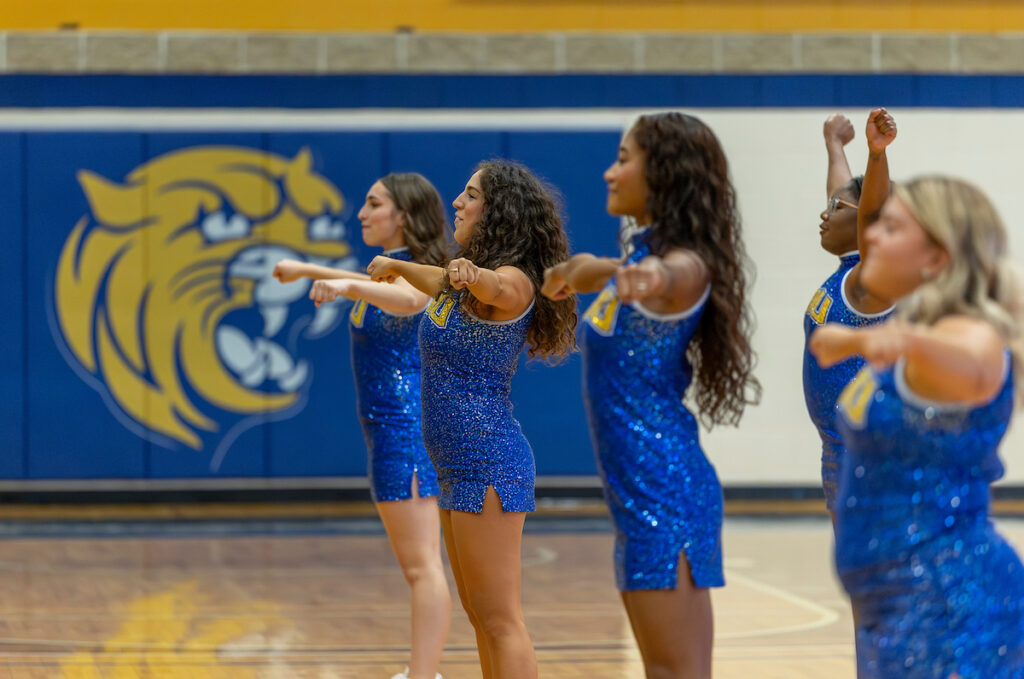 A group of dancers in blue sequined outfits perform synchronized movements on a gymnasium floor, with a large tiger mascot logo visible on the wall behind them.