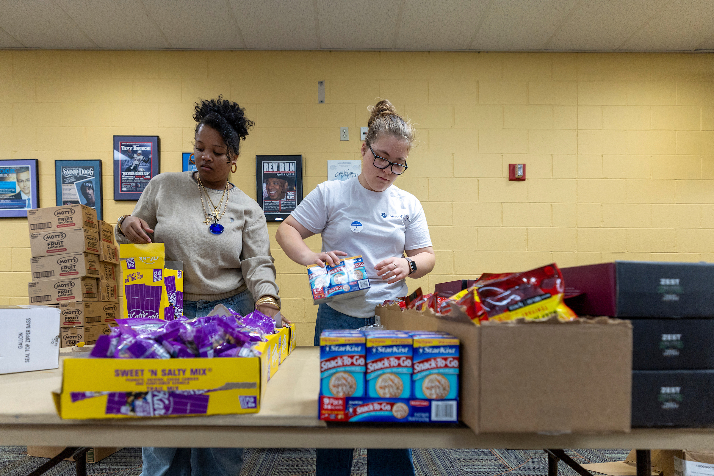 Two individuals sort and organize snack items on a table in a room with yellow walls.