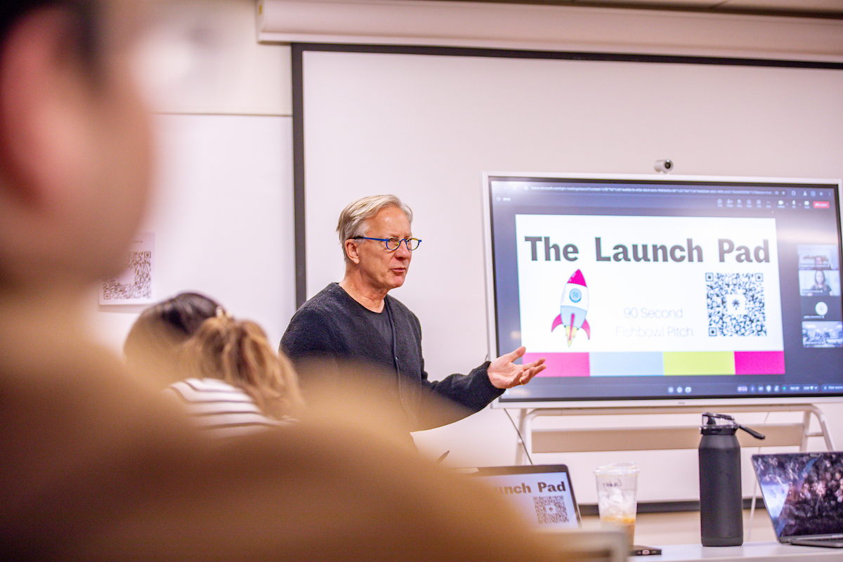 A person presents in front of an audience, gesturing toward a screen displaying "The Launch Pad" with a rocket graphic and QR code.