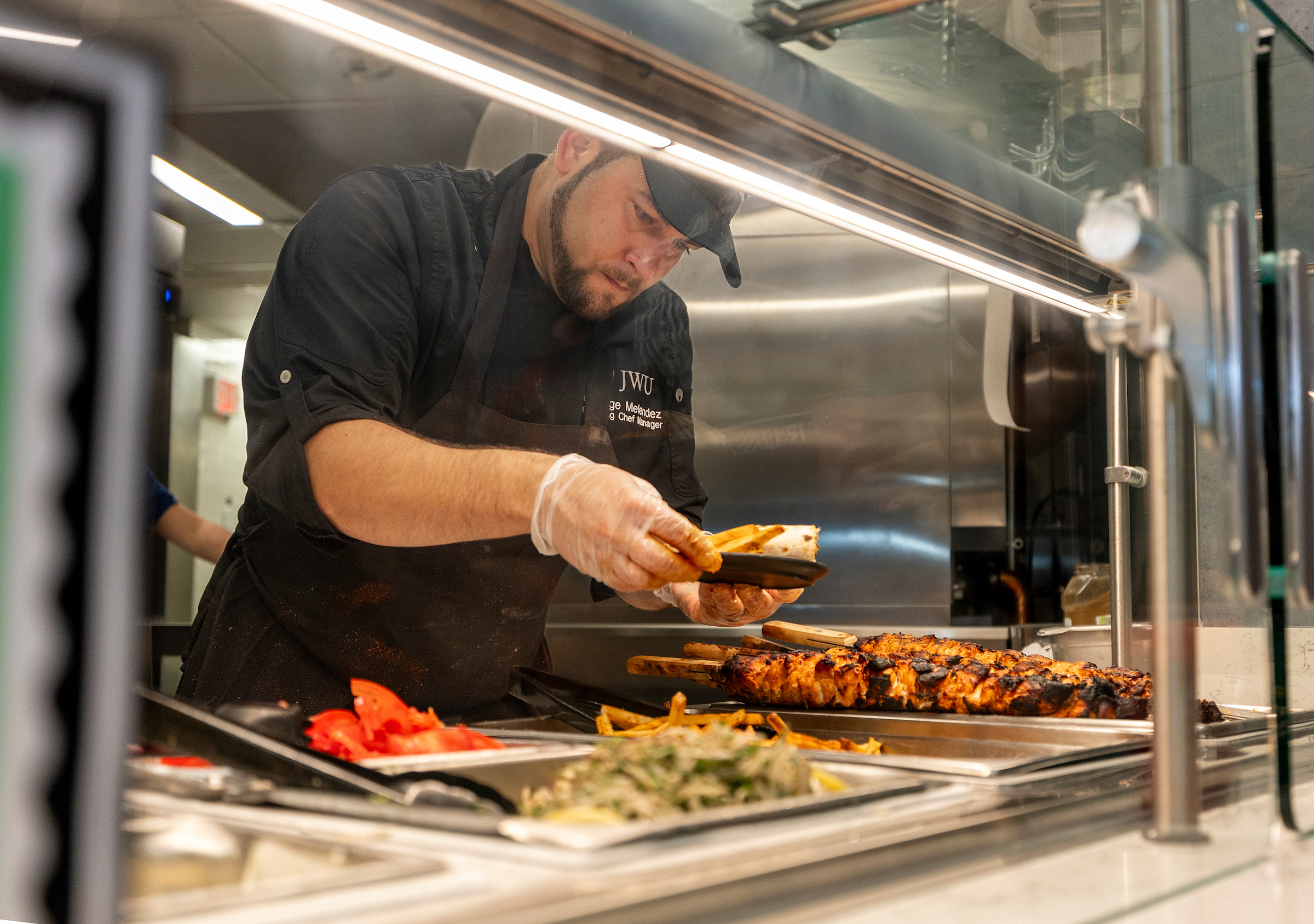 A chef prepares food behind a glass counter, assembling a dish with grilled meat and vegetables.