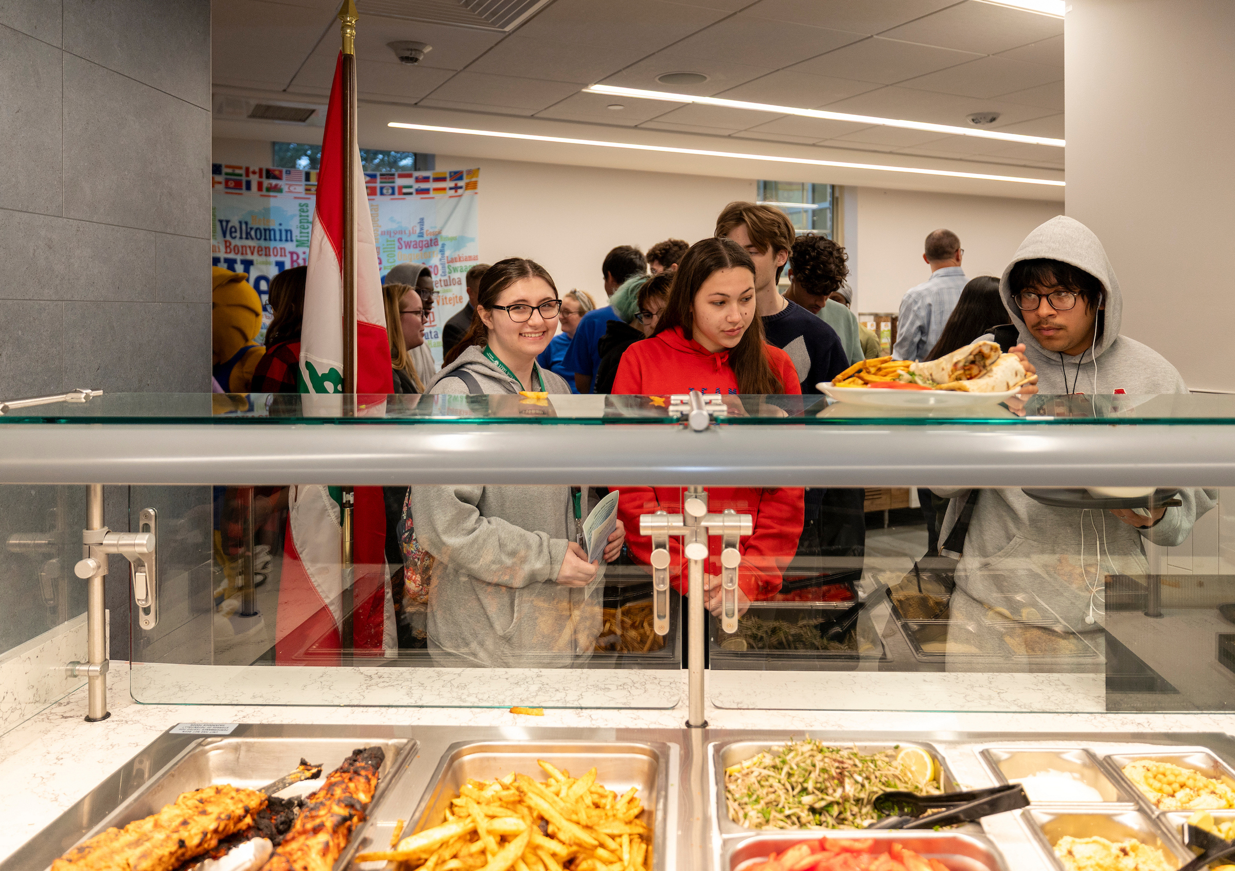 Students gather at a cafeteria counter, selecting food from trays while a flag and multilingual welcome signs are visible in the background.
