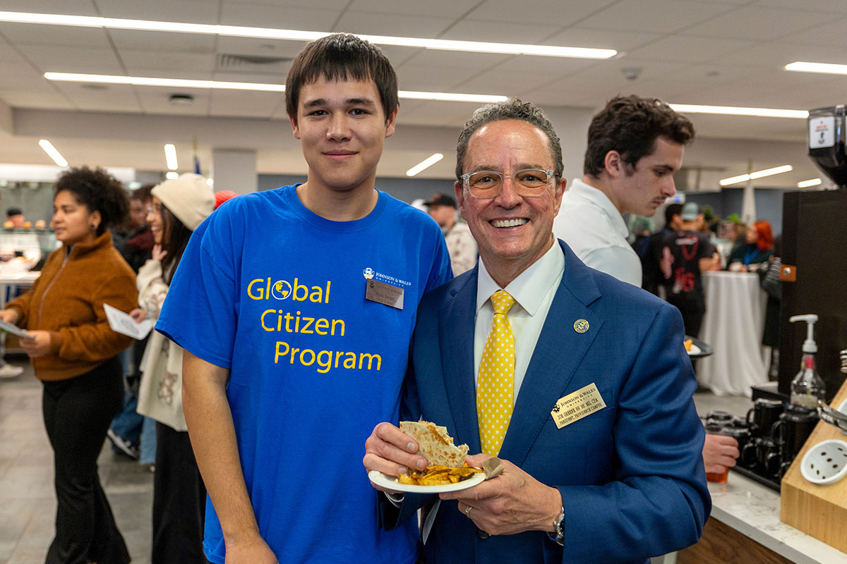 A young person in a Global Citizen Program shirt stands beside a formally dressed individual holding a plate of food at an indoor event.