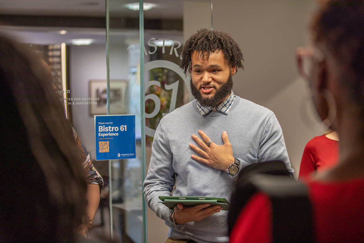 A person with curly hair and a beard, wearing a gray sweater over a collared shirt, holds a tablet and gestures with one hand while speaking to others in a modern indoor setting.