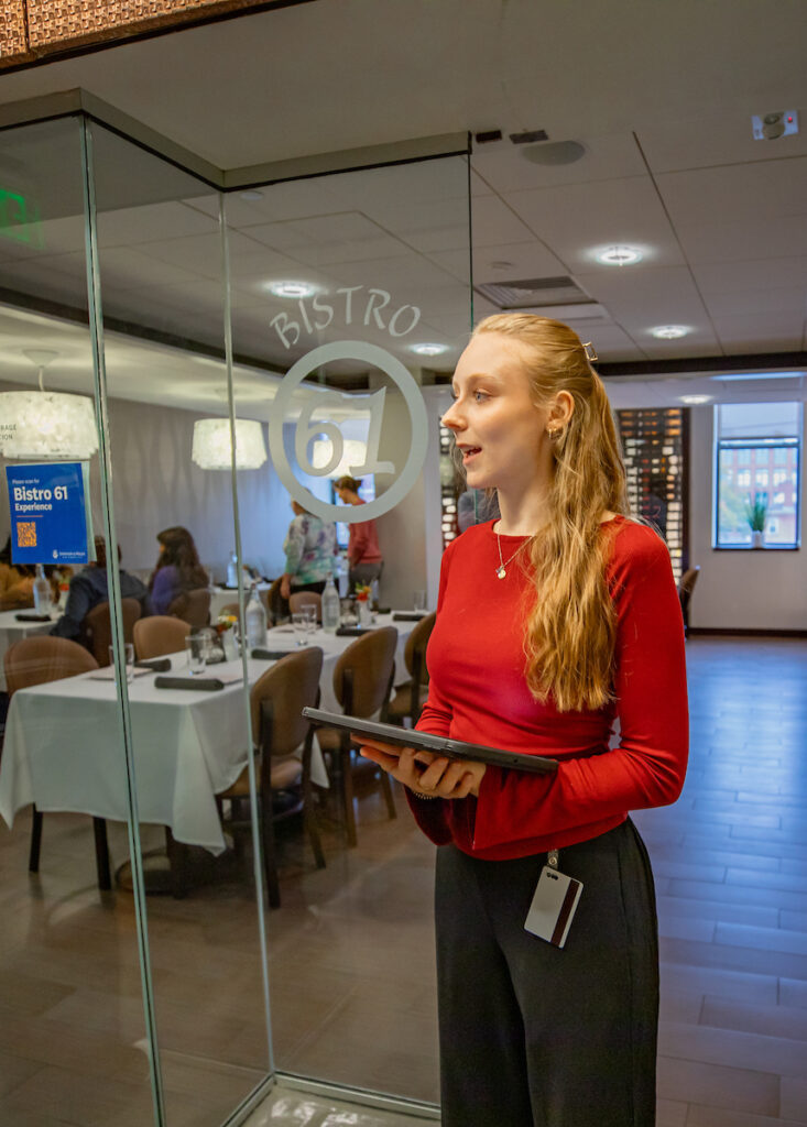 A server in a red top stands outside a glass door marked "Bistro 61," holding a tablet and smiling as guests dine inside.