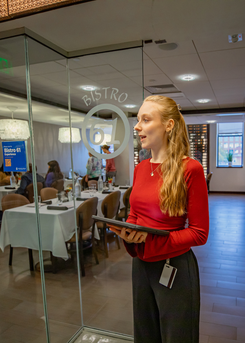 A server in a red top stands outside a glass door marked "Bistro 61," holding a tablet and smiling as guests dine inside.