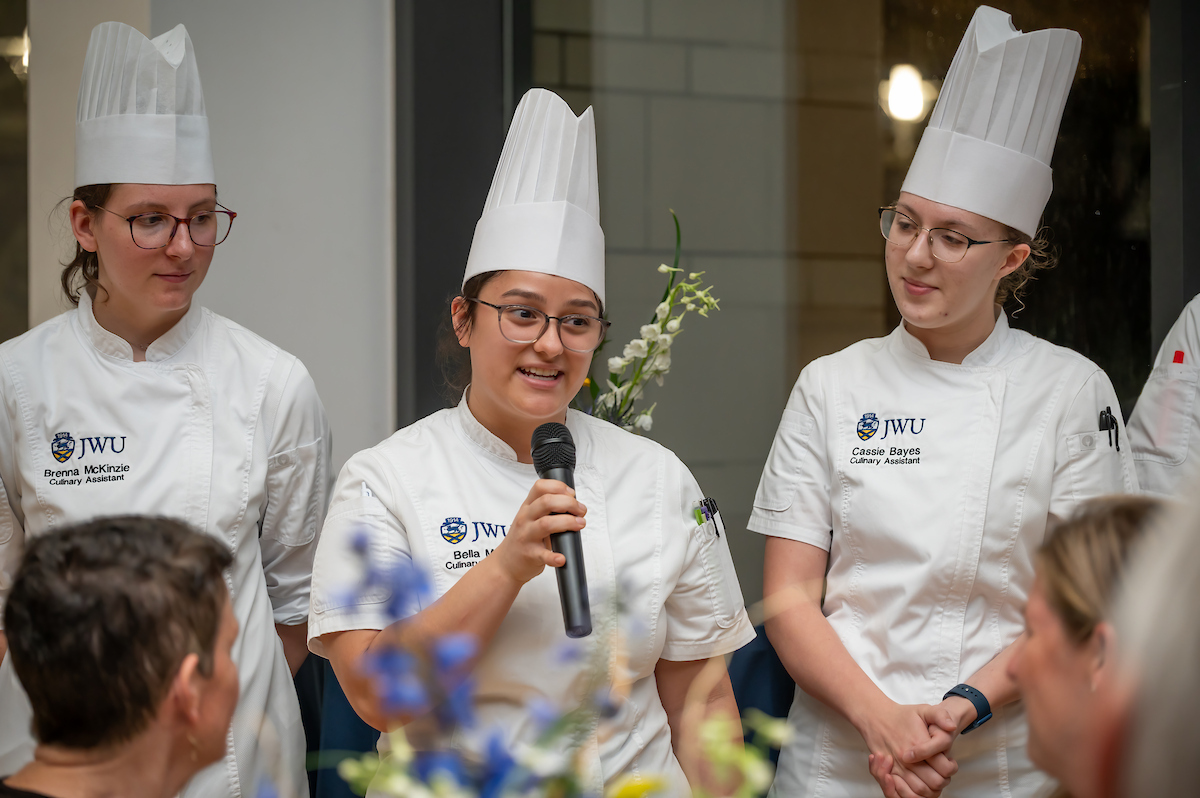 Three individuals in chef uniforms stand together, one speaking into a microphone while the others listen attentively.