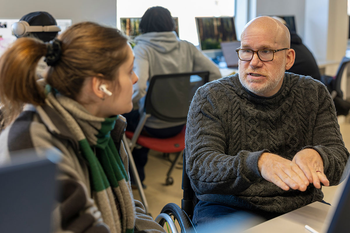 A professor engages in conversation with a student at a desk in a classroom.