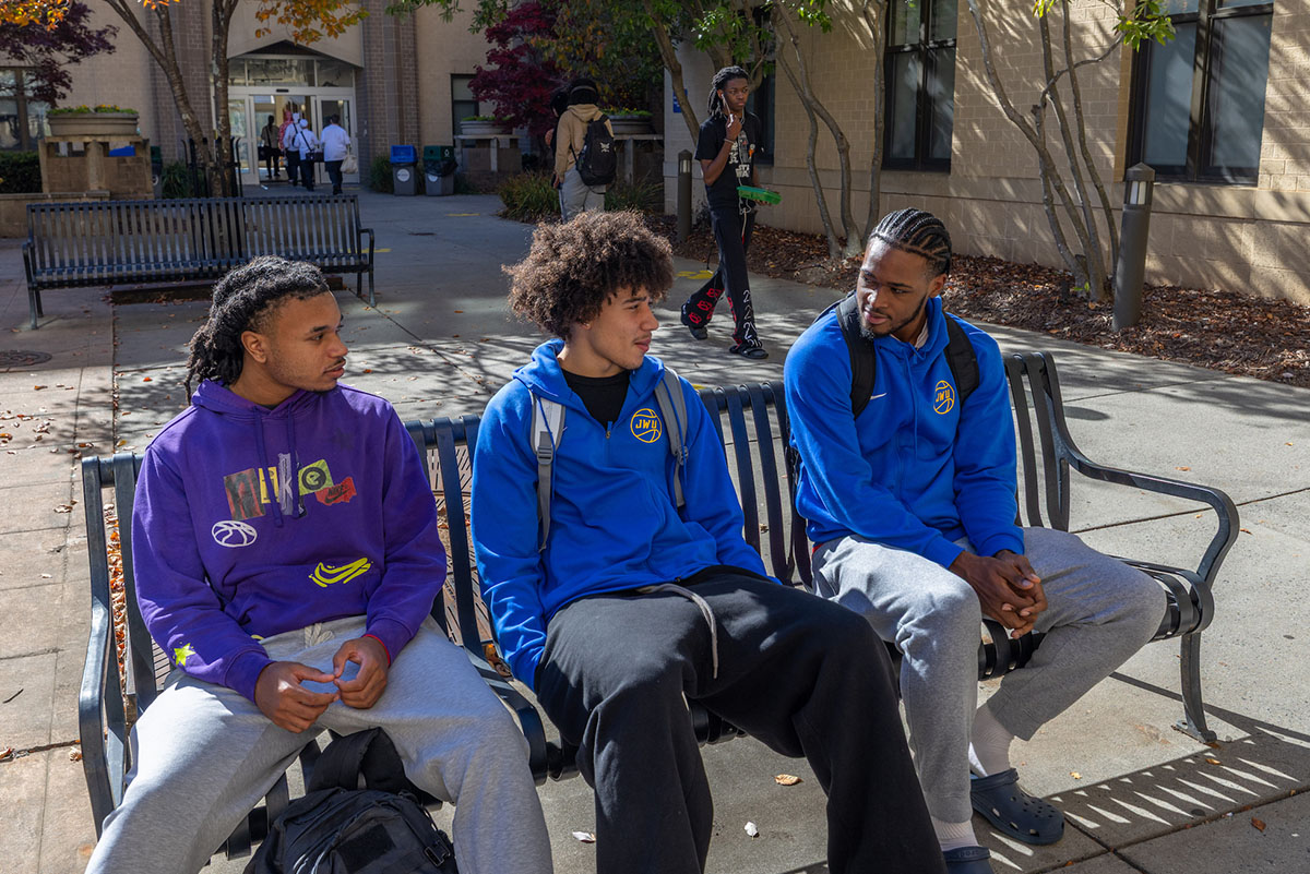 Three students sit on a bench outdoors, engaged in conversation, with autumn foliage and a building in the background.