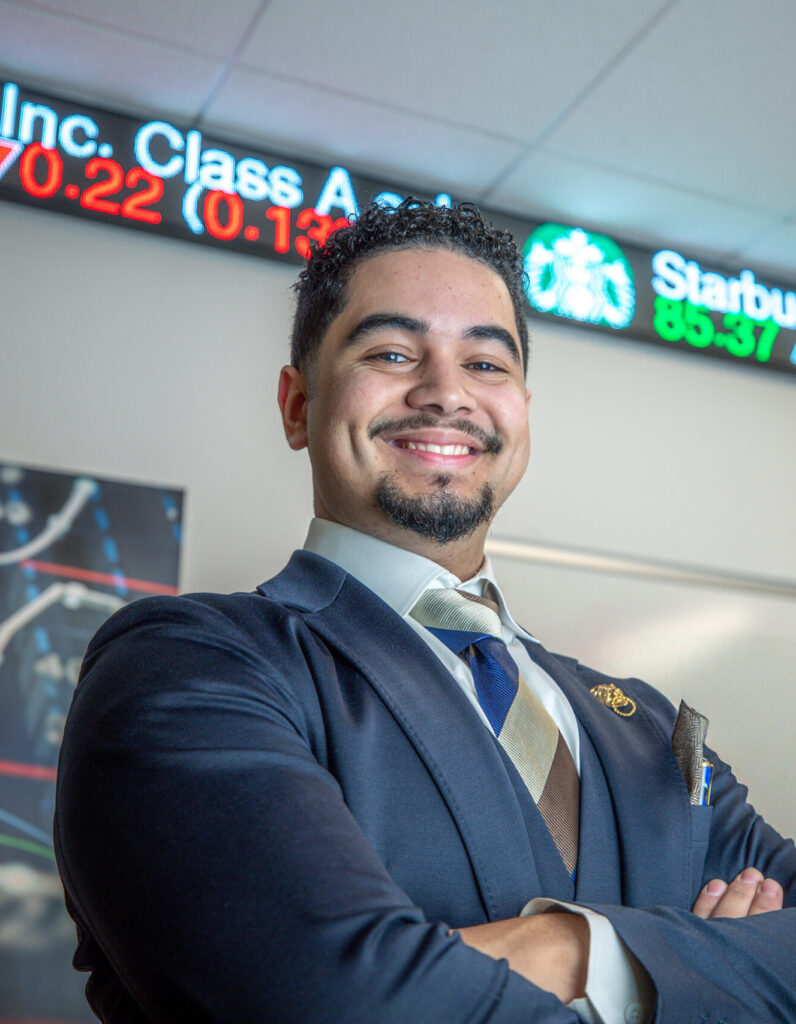A professionally dressed individual stands with arms crossed in front of a digital stock ticker displaying Shopify and Starbucks stock prices.