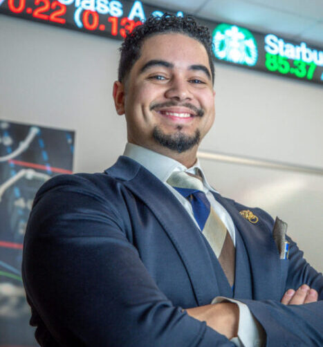 A professionally dressed individual stands with arms crossed in front of a digital stock ticker displaying Shopify and Starbucks stock prices.