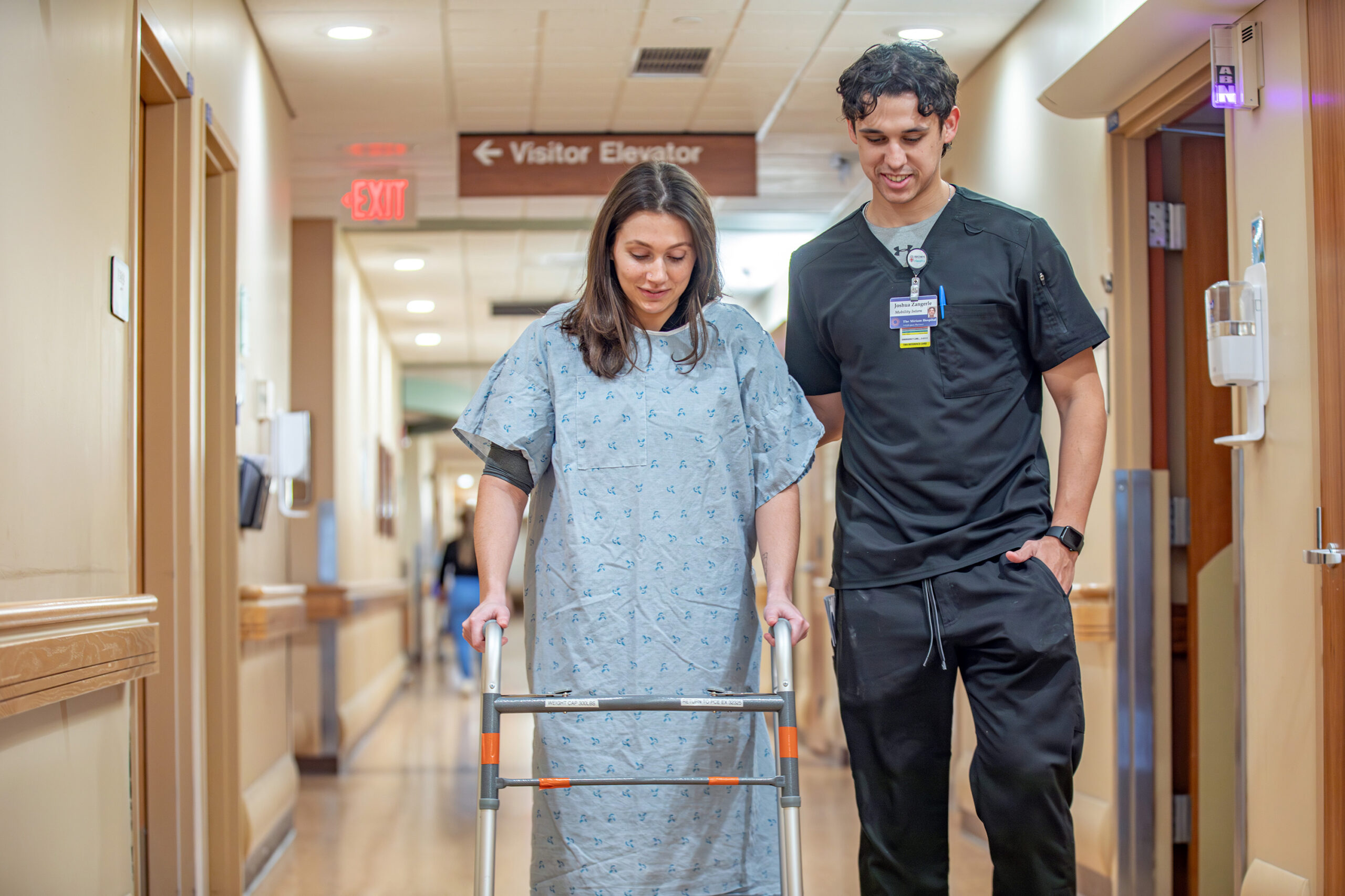 A person in a hospital gown uses a walker while being assisted by a healthcare worker in a hospital hallway.