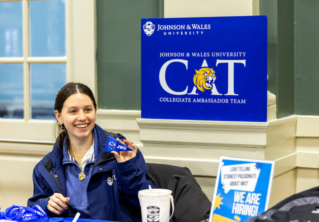 A smiling individual wearing a blue jacket sits at a table with a Johnson & Wales University CAT sign and a “We Are Hiring!” sign nearby.