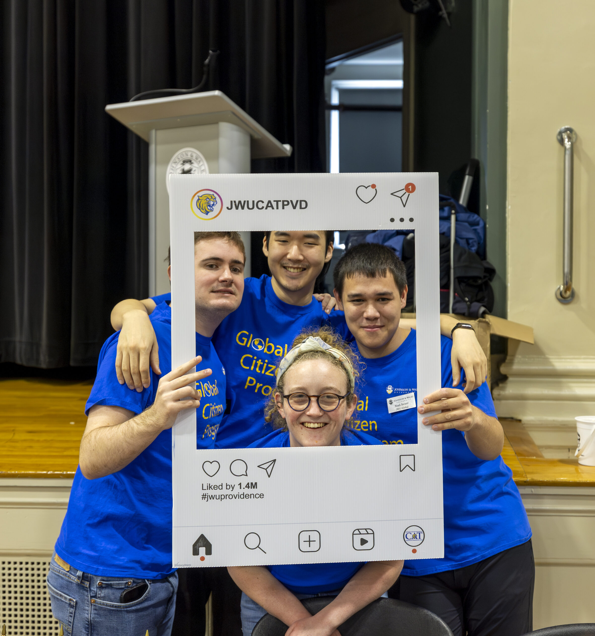 Four individuals in blue shirts pose together with a large Instagram-style photo frame, smiling for the camera at what appears to be an event.