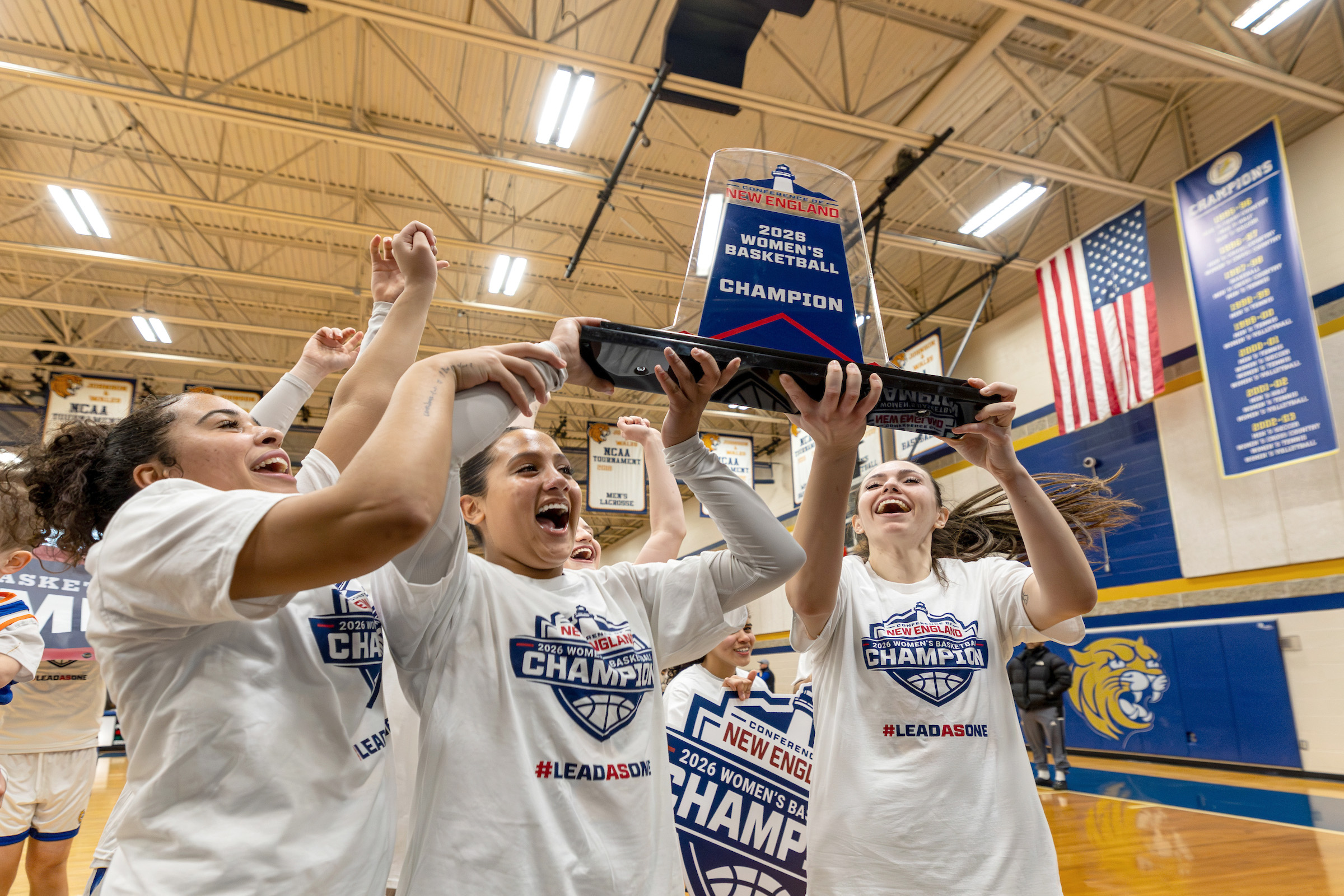 A group of athletes in matching championship shirts joyfully lift a trophy above their heads in a gymnasium.