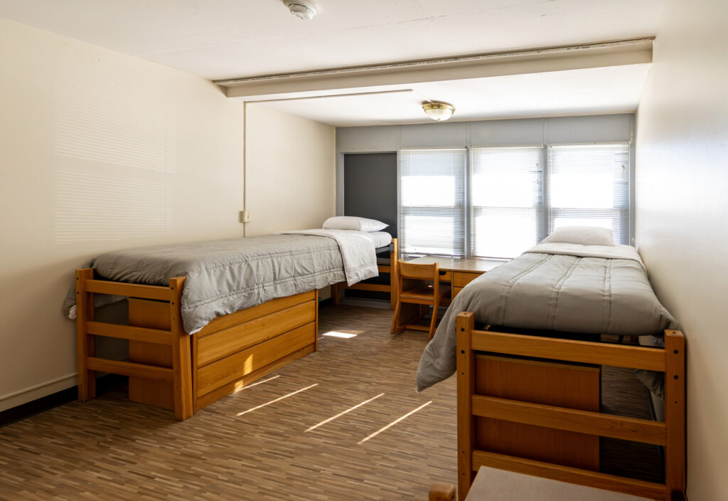 A clean, minimalist dorm room with two wooden bunk beds, a desk, and windows with blinds.