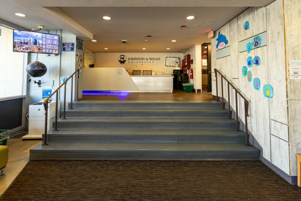 The entrance to Johnson & Wales University features a reception desk, stairs leading up to it, and colorful handprint decorations on the wall.