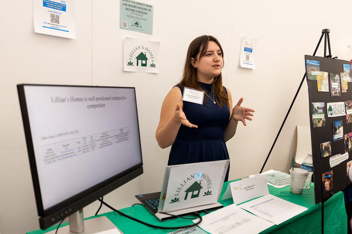 A person stands at a booth for Lillian's Homes, gesturing while speaking, with a monitor displaying data and promotional materials on a green-covered table.