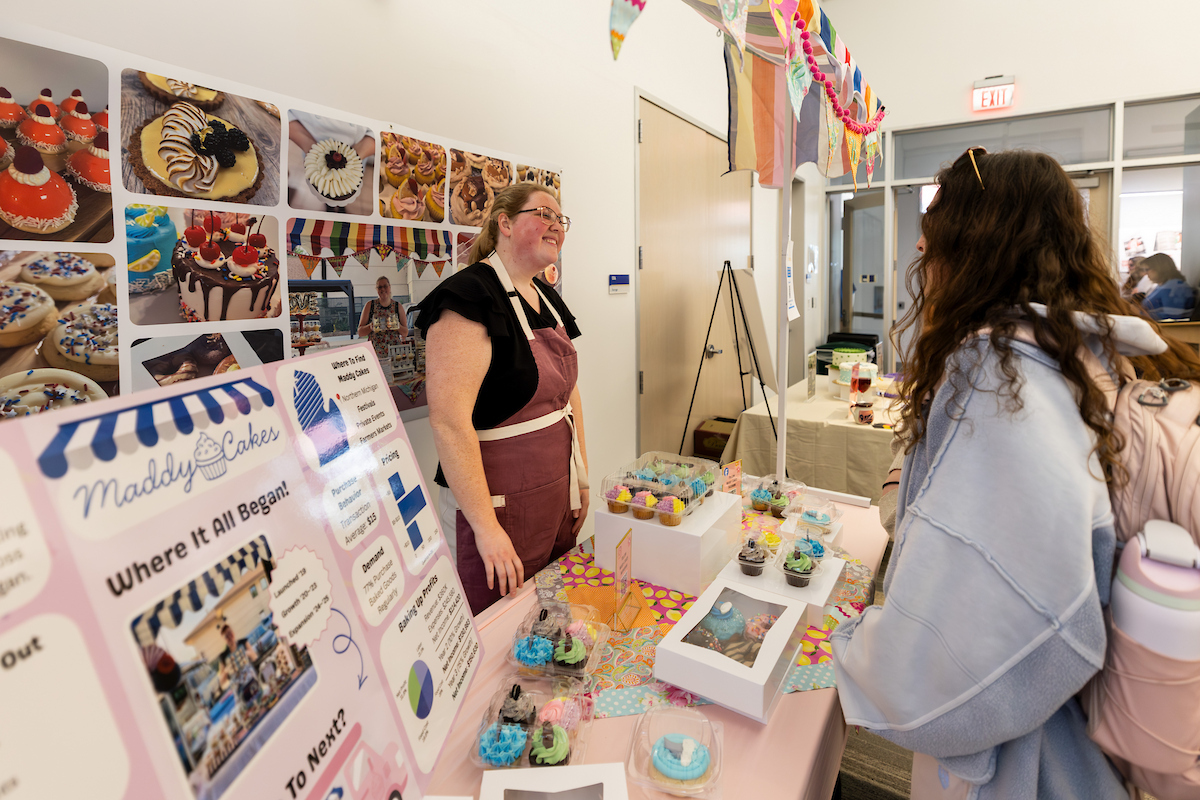 A person in an apron stands behind a table displaying cupcakes and promotional materials, engaging with a customer at what appears to be a bake sale or market stall.