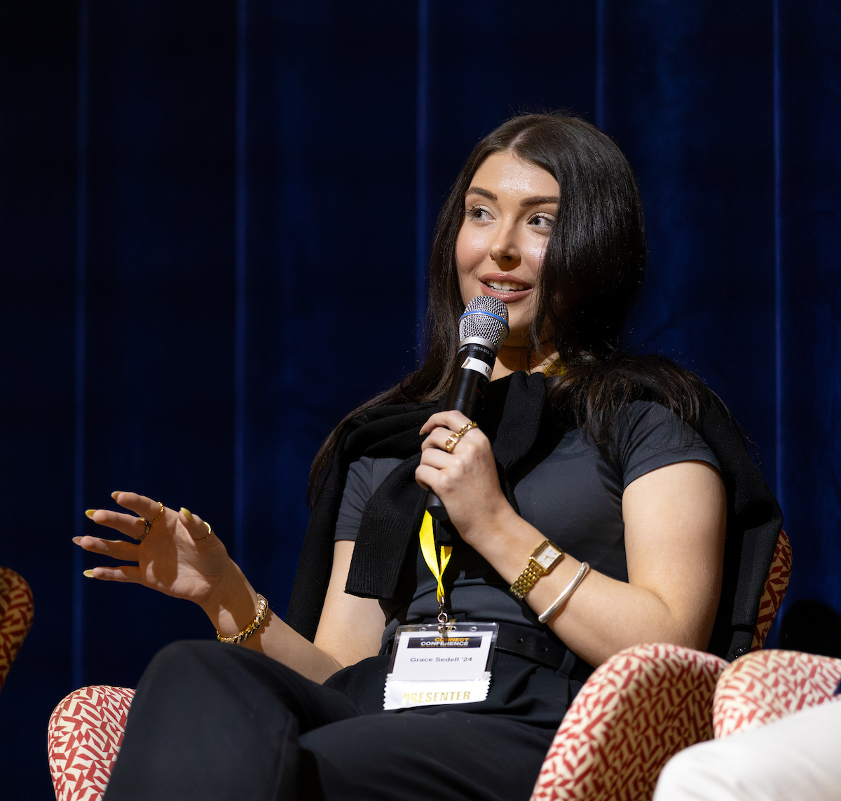 A person speaks into a microphone while seated on a patterned chair, wearing a lanyard with a name tag, against a dark blue backdrop.