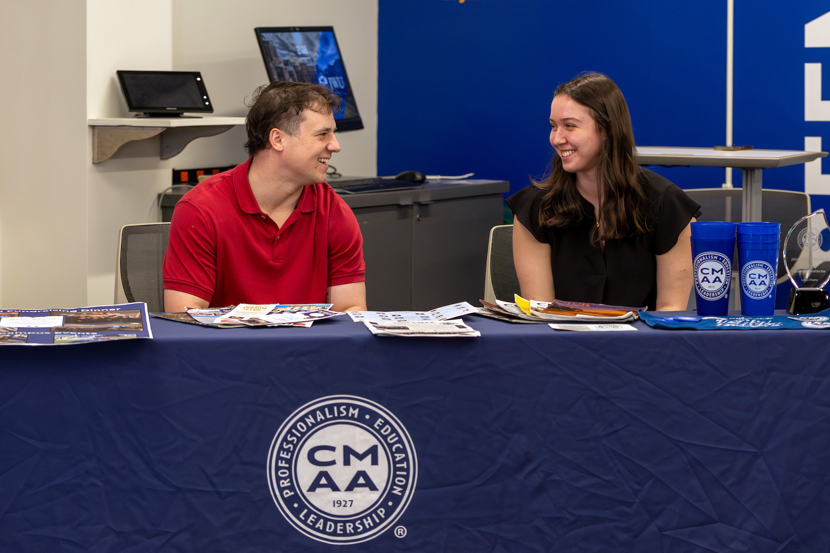 Two individuals engage in conversation at a table covered with a Johnson and Wales University Rhode Island Chapter banner, surrounded by promotional materials and branded items.