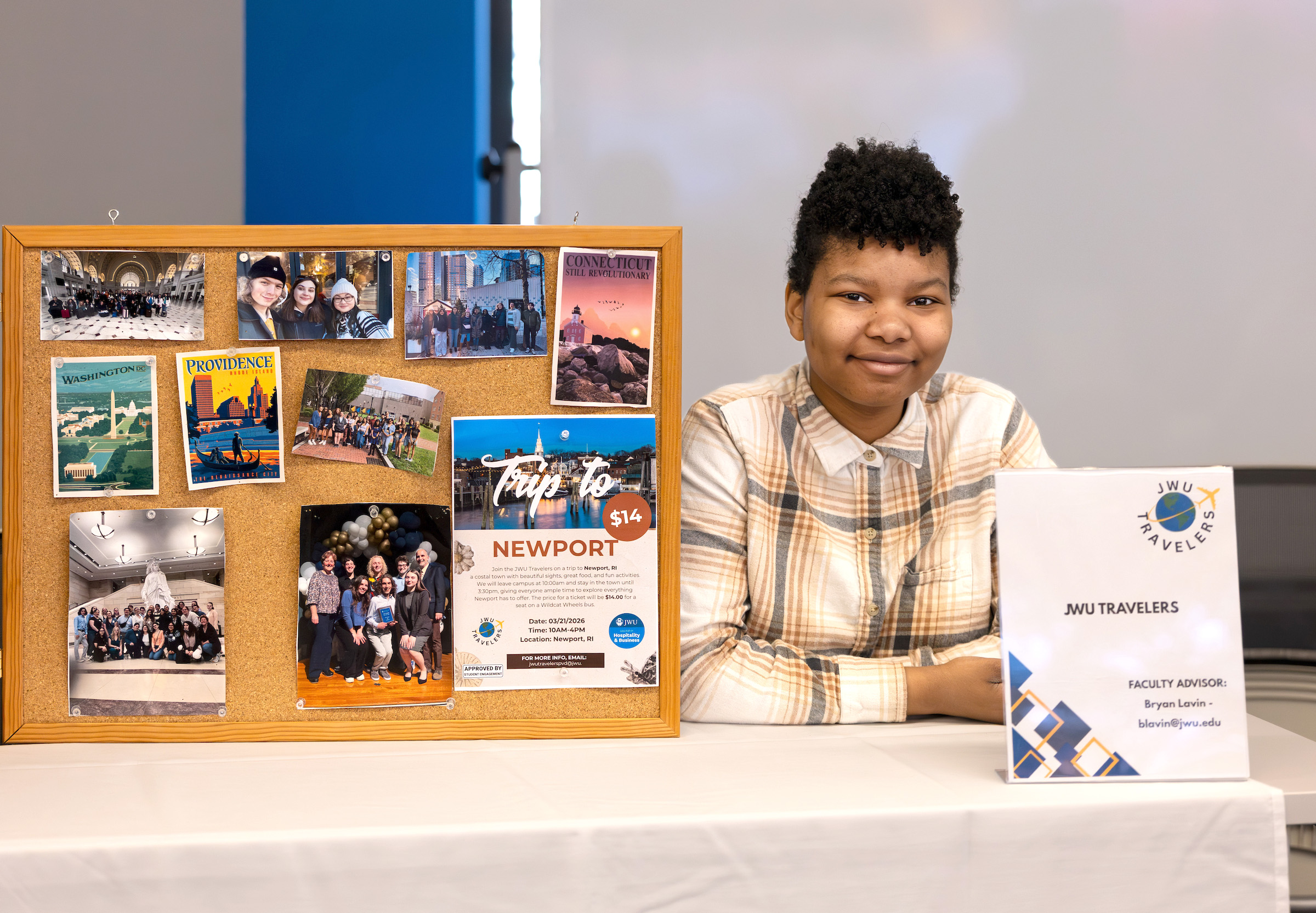 A young person smiles while seated at a table beside a display board featuring travel photos and a “Trip to Newport” poster, alongside a JWU Travelers program sign.
