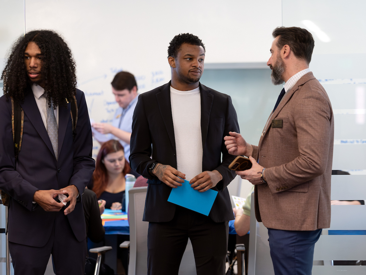 Three individuals in professional attire stand in an office setting, engaged in conversation, with one holding a blue folder and another holding a phone.