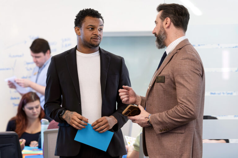 Three individuals in professional attire stand in an office setting, engaged in conversation, with one holding a blue folder and another holding a phone.