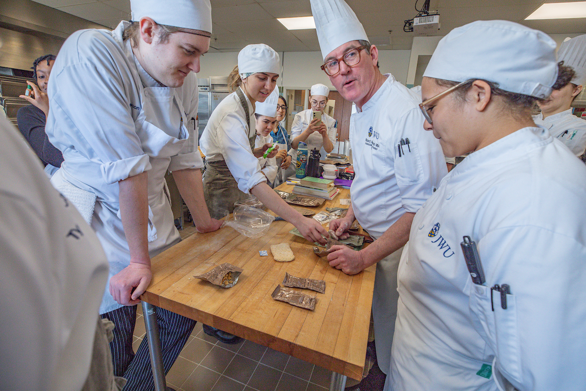 Several individuals in chef attire gather around a wooden table in a kitchen, attentively examining food items and ingredients.