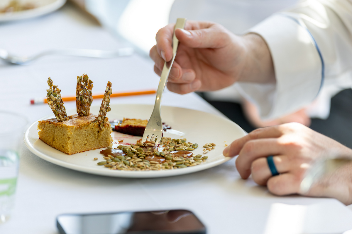 A person uses a fork to eat a dessert featuring a square cake topped with seed-crusted sticks and a pile of seeds and syrup on a white plate.
