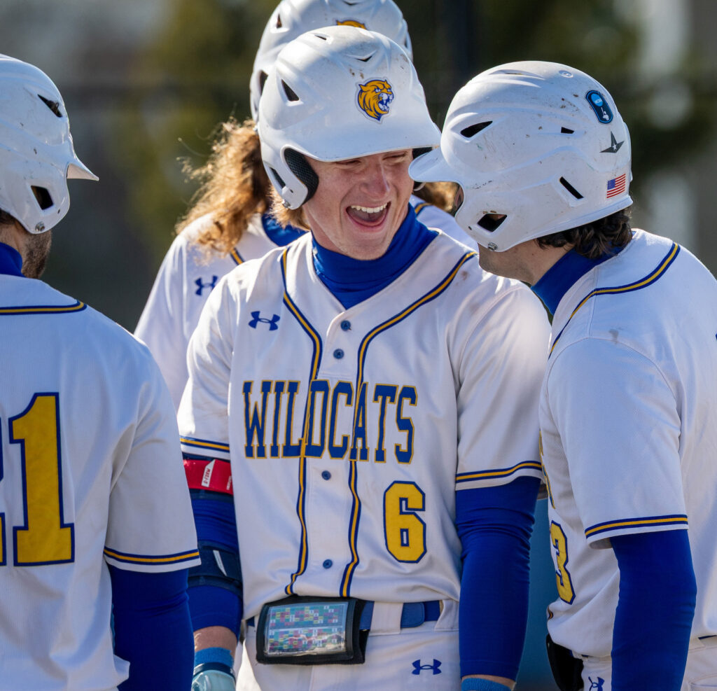 A group of baseball players in white and blue uniforms with "WILDCATS" and the number 6, celebrate on the field.