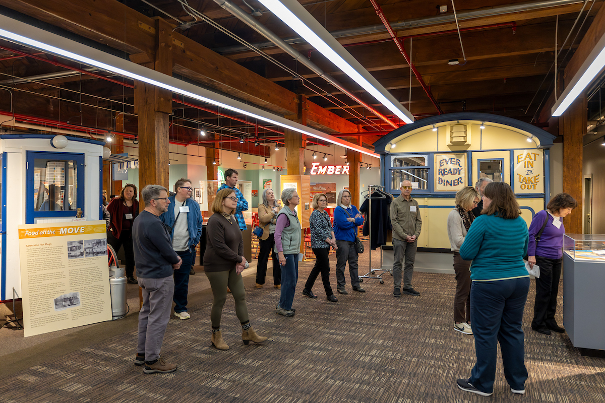 A group of visitors explores a museum exhibit featuring vintage food service booths, including a replica diner and a blue service station, under exposed wooden beams and industrial lighting.