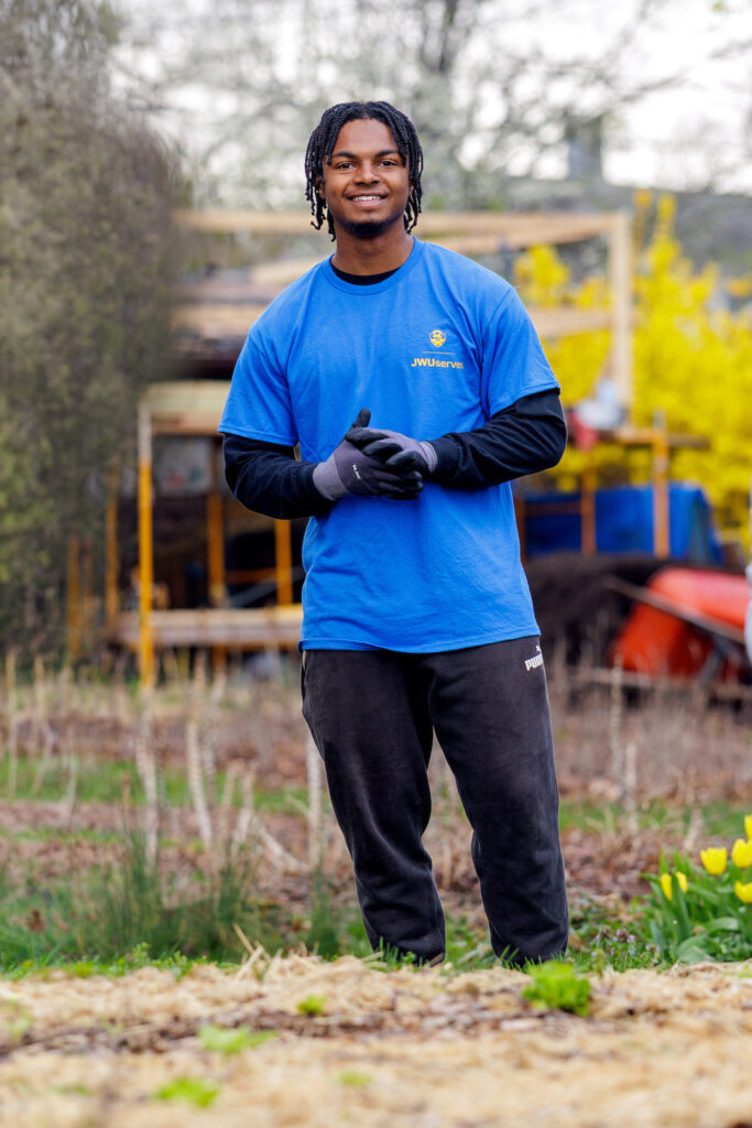 A person in a blue JWUserve shirt stands smiling in a garden with tulips, while others are visible in the background.
