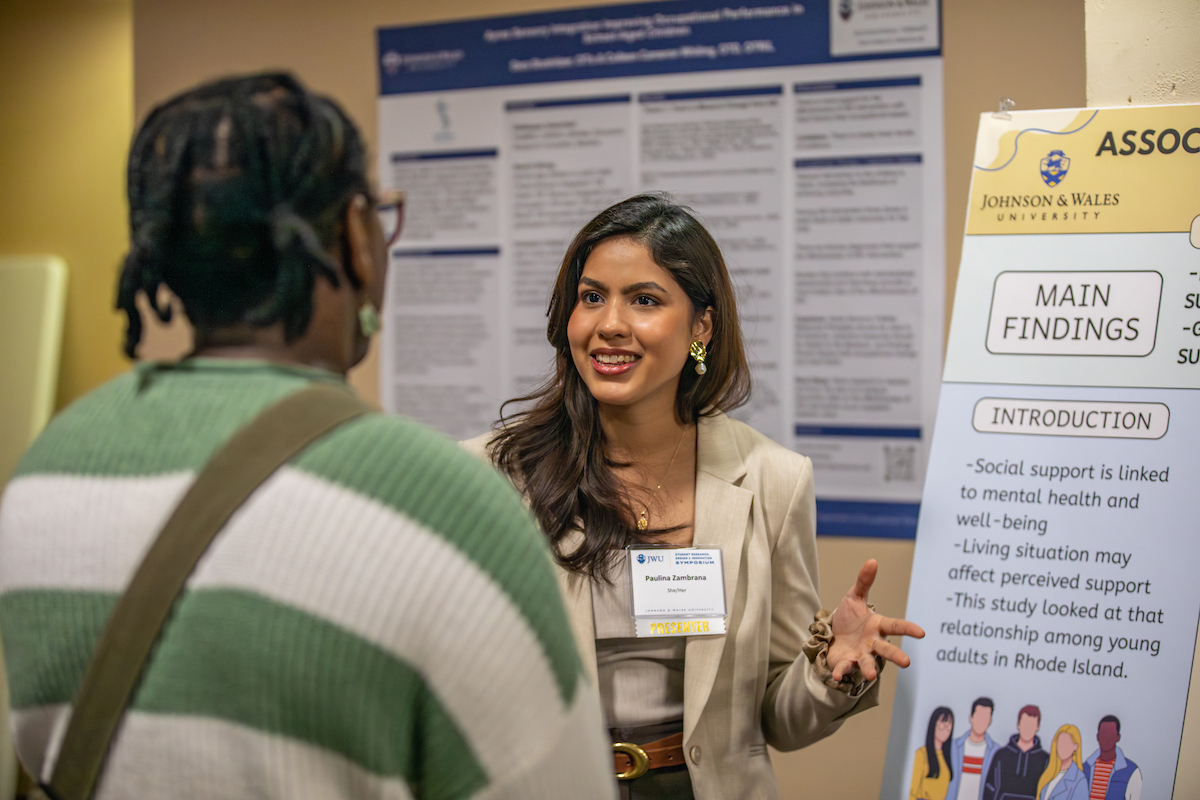 A person in a beige blazer gestures while speaking to another individual at an academic event, with research posters from Johnson 